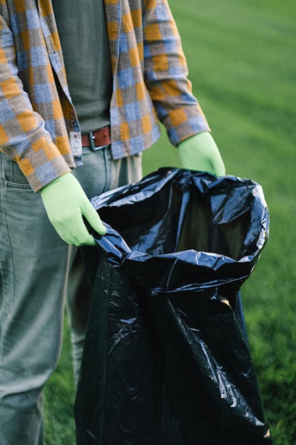 A person wearing a checkered shirt and green gloves is holding open a large black plastic rubbish bag, preparing for waste disposal. The individual is standing outdoors on a grassy area, with no visib